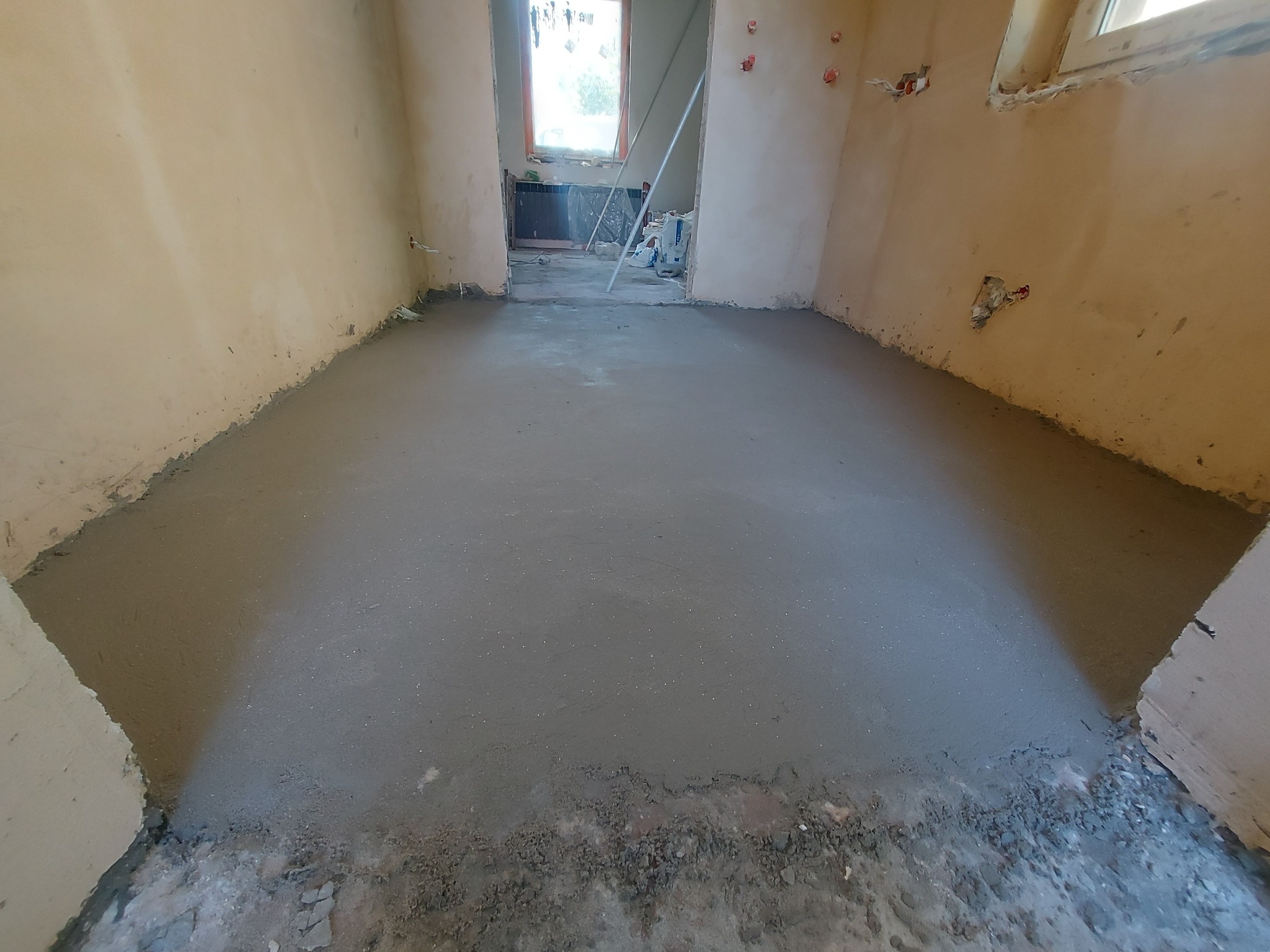 Wyrównana, szara wylewka podłogowa w trakcie schnięcia, widoczne niedoskonałości ścian i wystające przewody elektryczne, w tle okno z widokiem na zieleń.