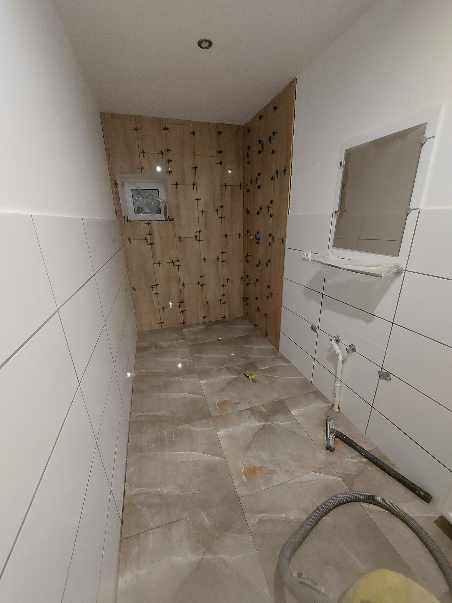 Łazienka w trakcie wykańczania: częściowo ułożone płytki imitujące drewno na ścianie, białe płytki na pozostałych ścianach, marmurowe płytki podłogowe, narzędzia glazurnicze na podłodze.