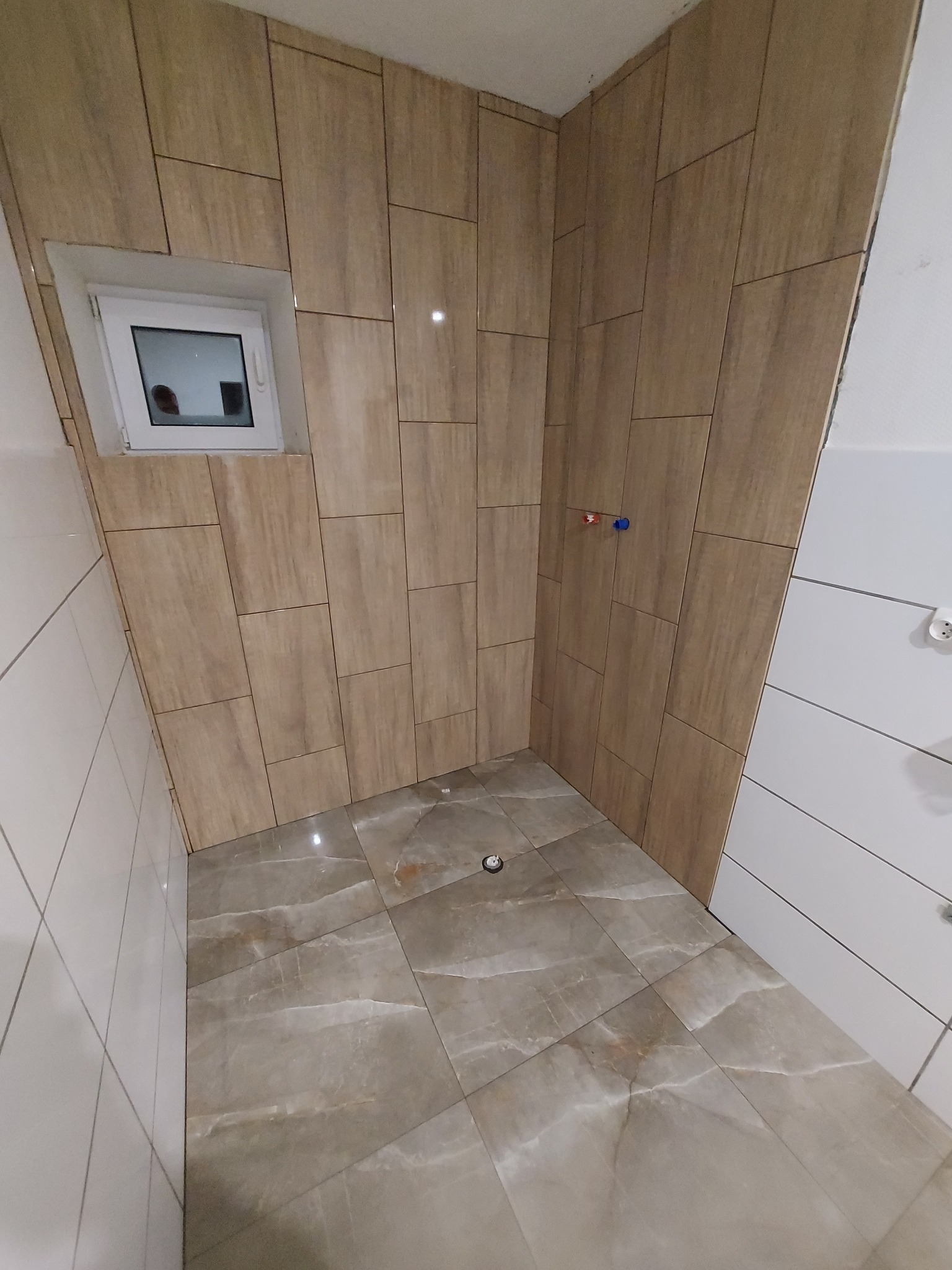 Wykończona łazienka z beżowymi płytkami imitującymi drewno na ścianach i marmurową podłogą, widoczne małe okno i instalacja wodna z czerwonym i niebieskim zaworem.