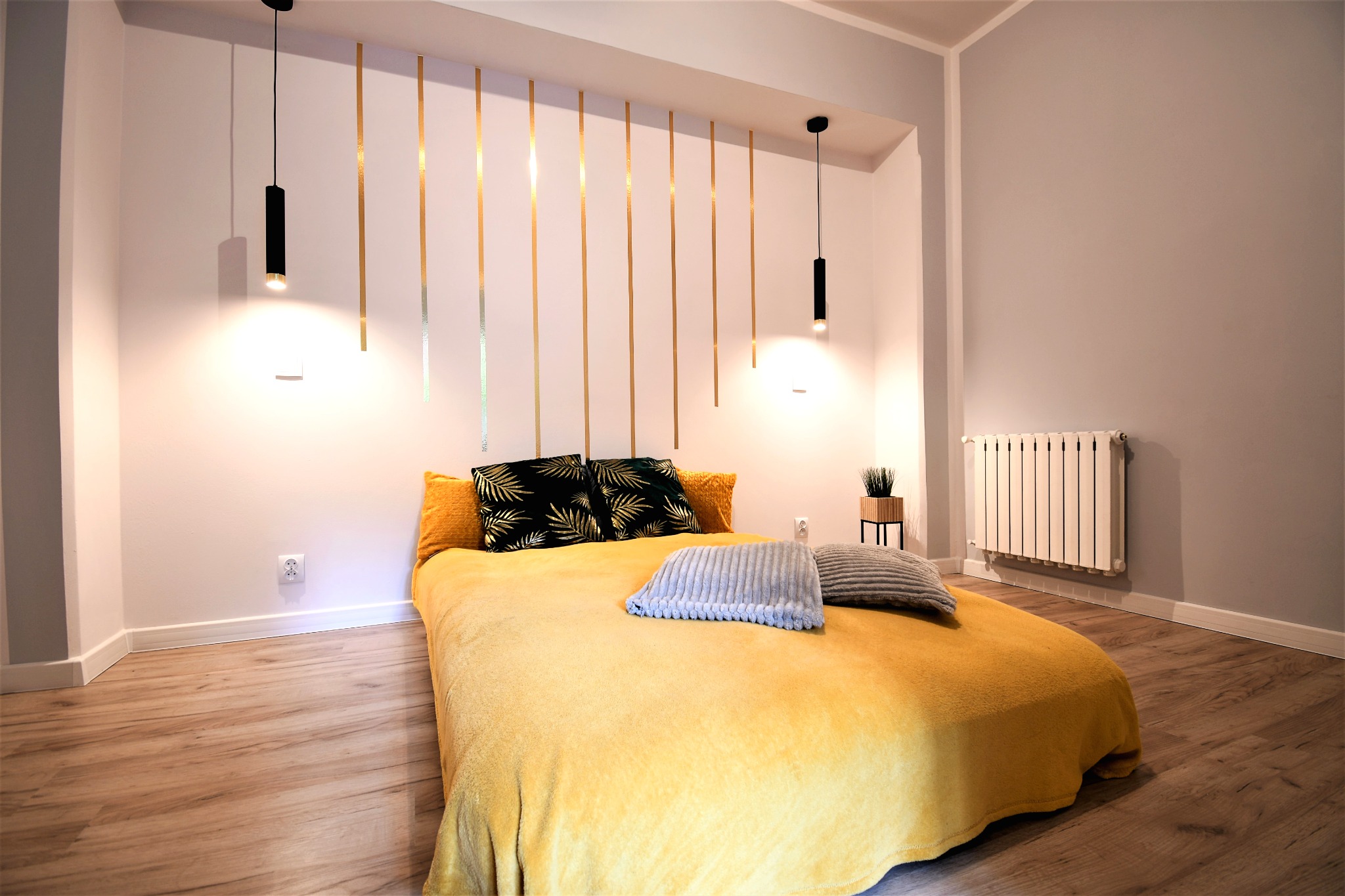 Jasna sypialnia z drewnianą podłogą, żółtą narzutą na łóżku, czarnymi lampami wiszącymi i dekoracyjnymi złotymi pasami na ścianie za wezgłowiem.