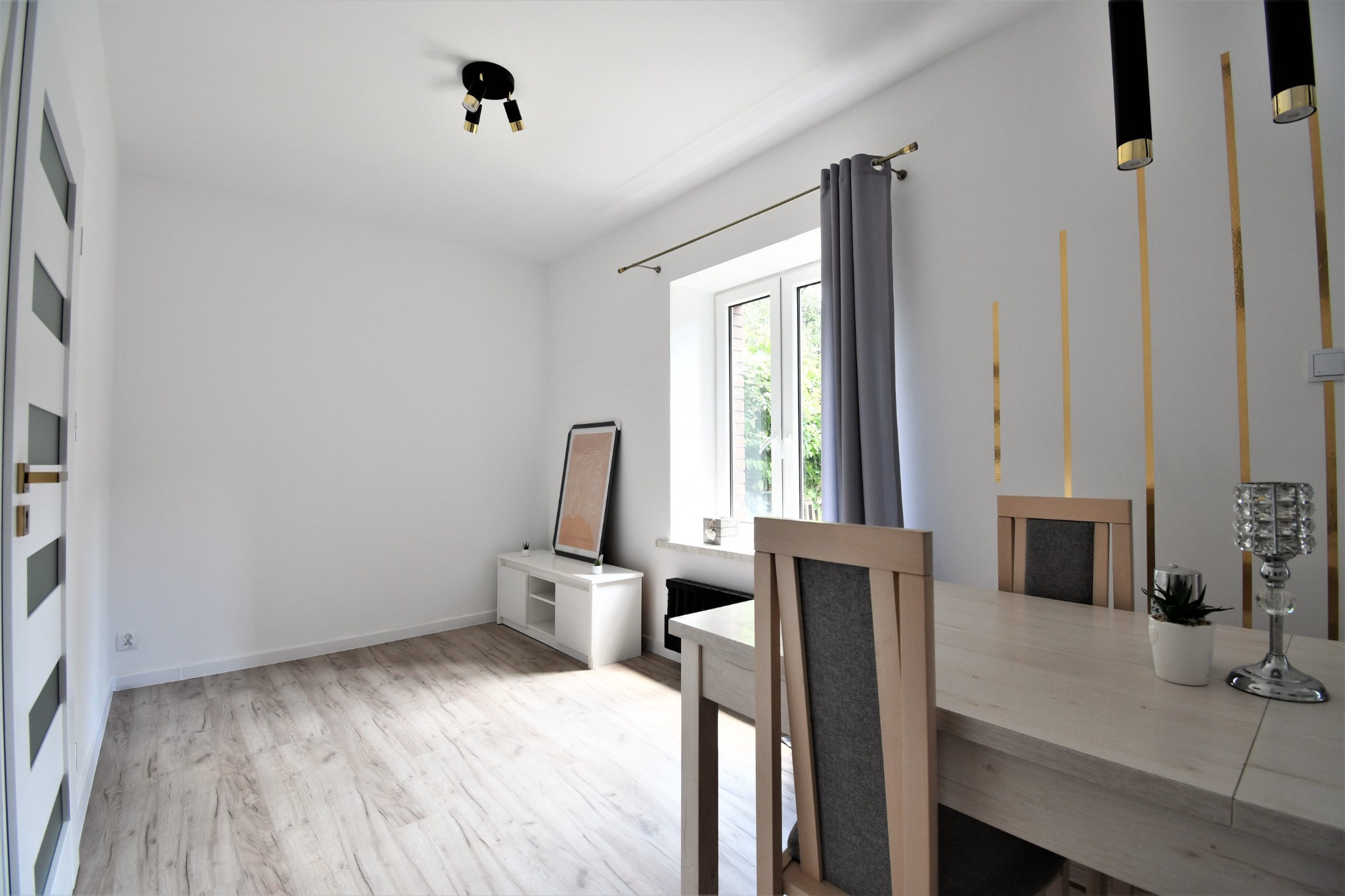 Jasne wnętrze jadalni z drewnianą podłogą, białymi ścianami z dekoracyjnymi złotymi paskami, drewnianym stołem z ozdobami i krzesłami z szarą tapicerką, oraz białymi meblami pod oknem.