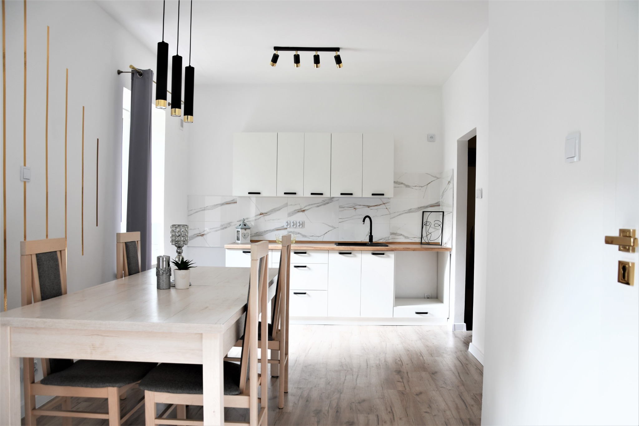 Jasna kuchnia z białymi szafkami, marmurowym backsplashiem i drewnianym blatem, uzupełniona o czarną armaturę i lampy, widoczny stół z krzesłami i dekoracyjnymi złotymi listwami na ścianie.