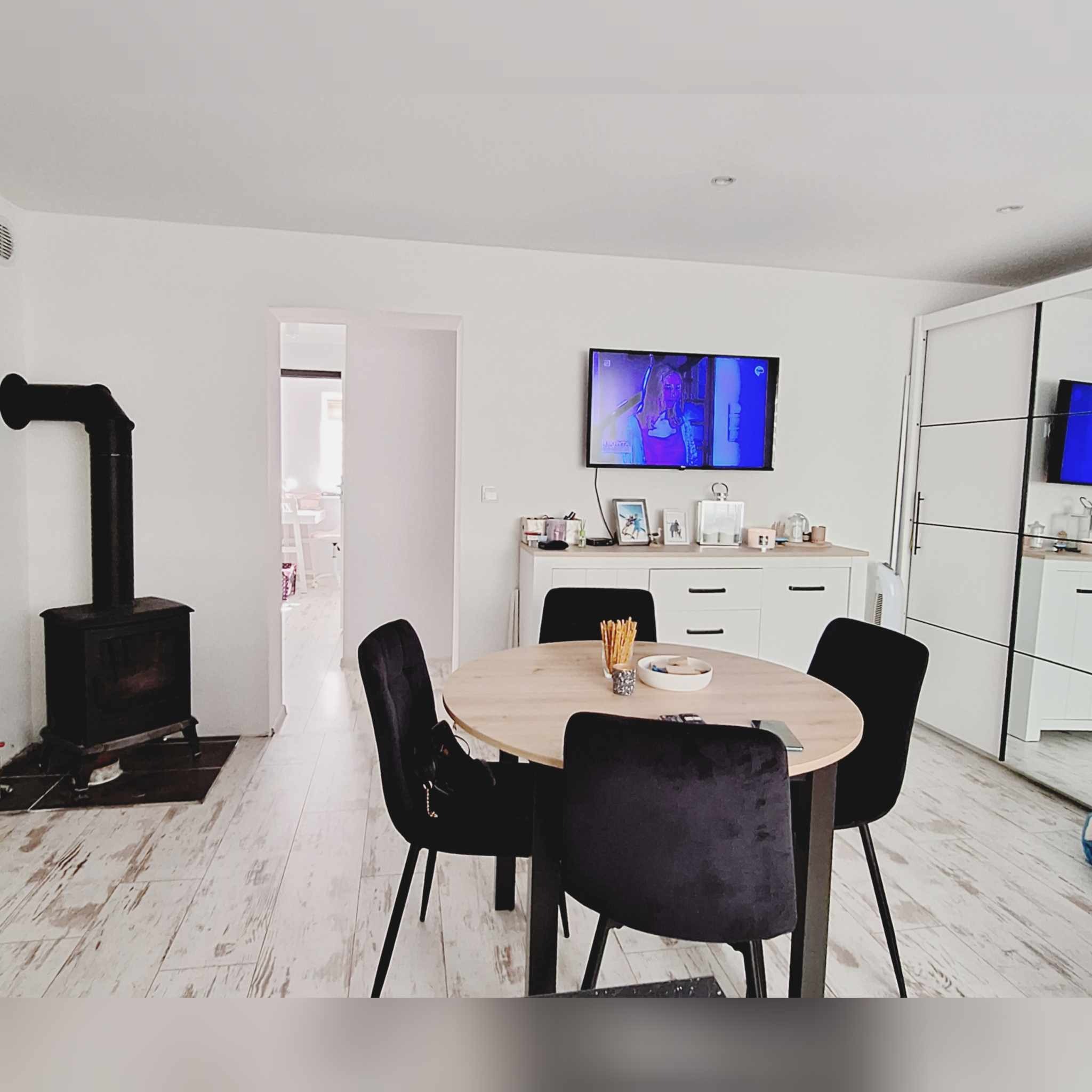 Jasne wnętrze pokoju z drewnianym stołem otoczonym czarnymi krzesłami, piecem kaflowym i telewizorem na ścianie