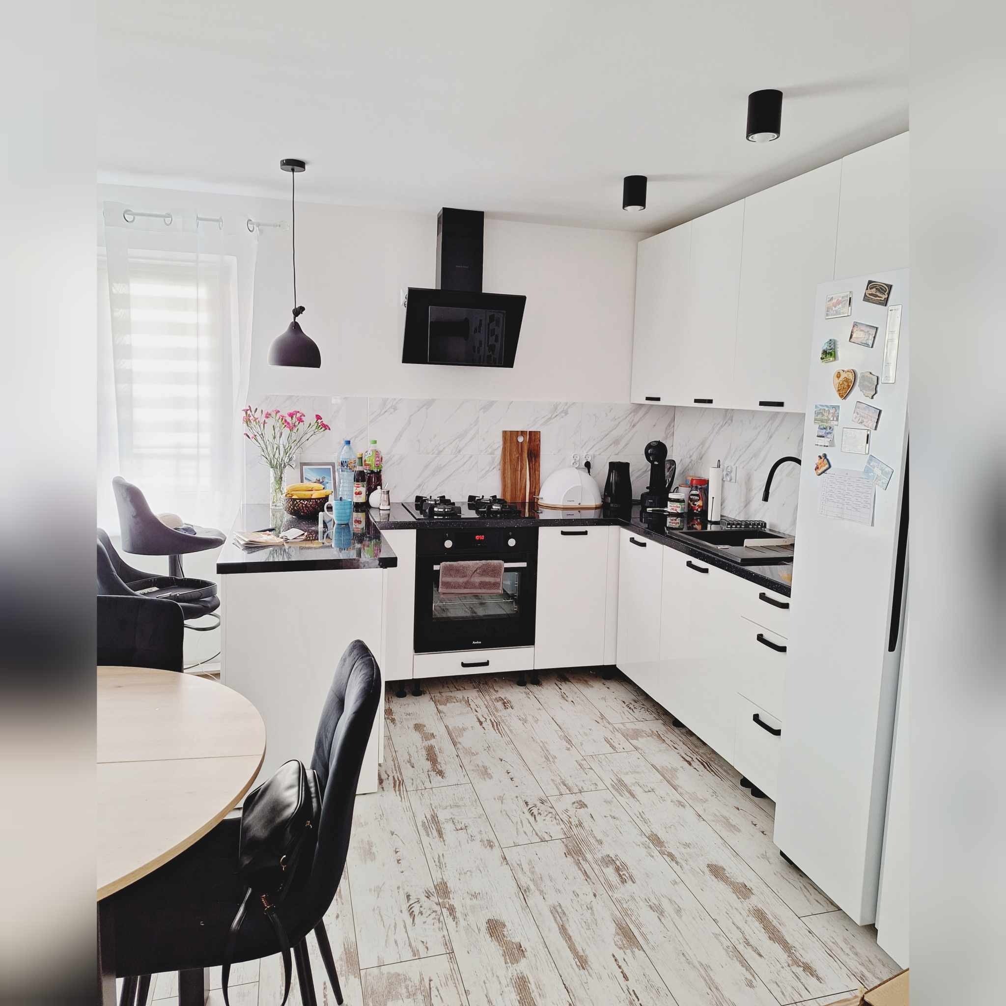 Jasna kuchnia z białymi szafkami, czarnym blatem i sprzętem AGD, drewniana podłoga z efektem postarzenia, dekoracje i kwiaty na blacie, nowoczesne oświetlenie.