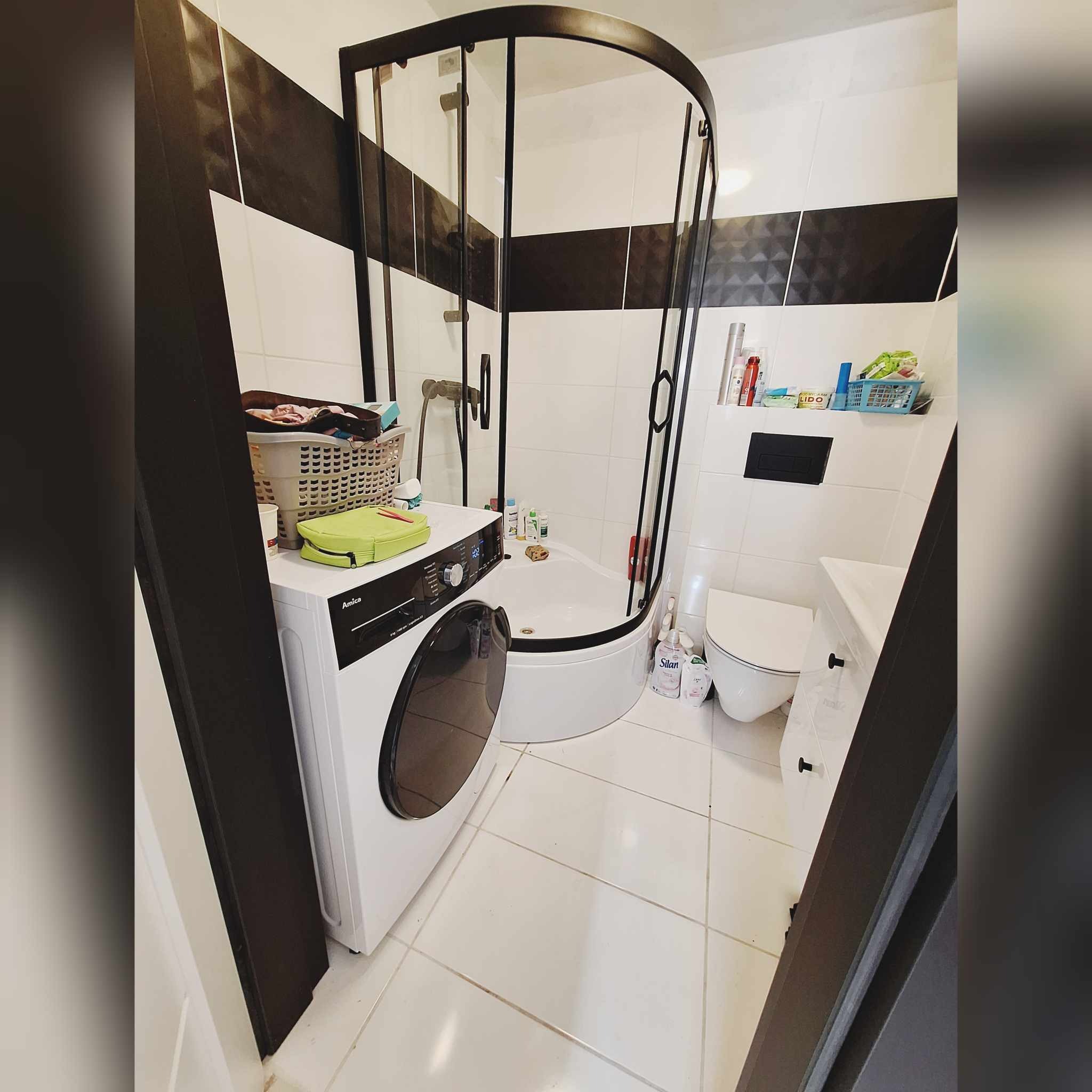 Wnętrze łazienki z narożną kabiną prysznicową z czarnymi ramami, białą pralką Amica z koszem na pranie i zieloną kosmetyczką na górze, białą toaletą i umywalką oraz czarno-białymi płytkami...