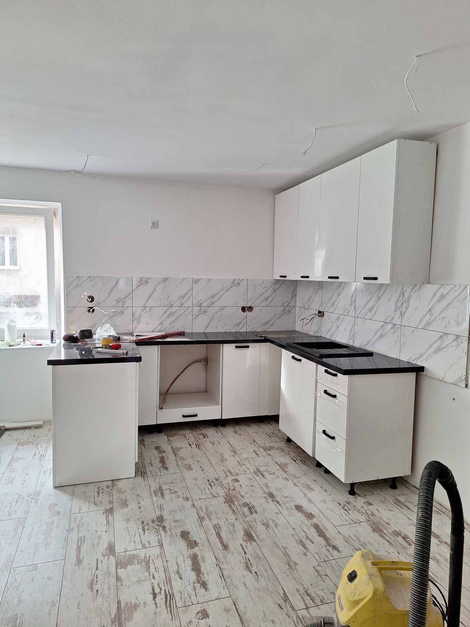 Nowa, biała kuchnia z czarnym blatem i marmurowymi płytkami na ścianie, w trakcie montażu, widoczny odkurzacz i narzędzia na blacie, surowe ściany z wystającymi kablami.