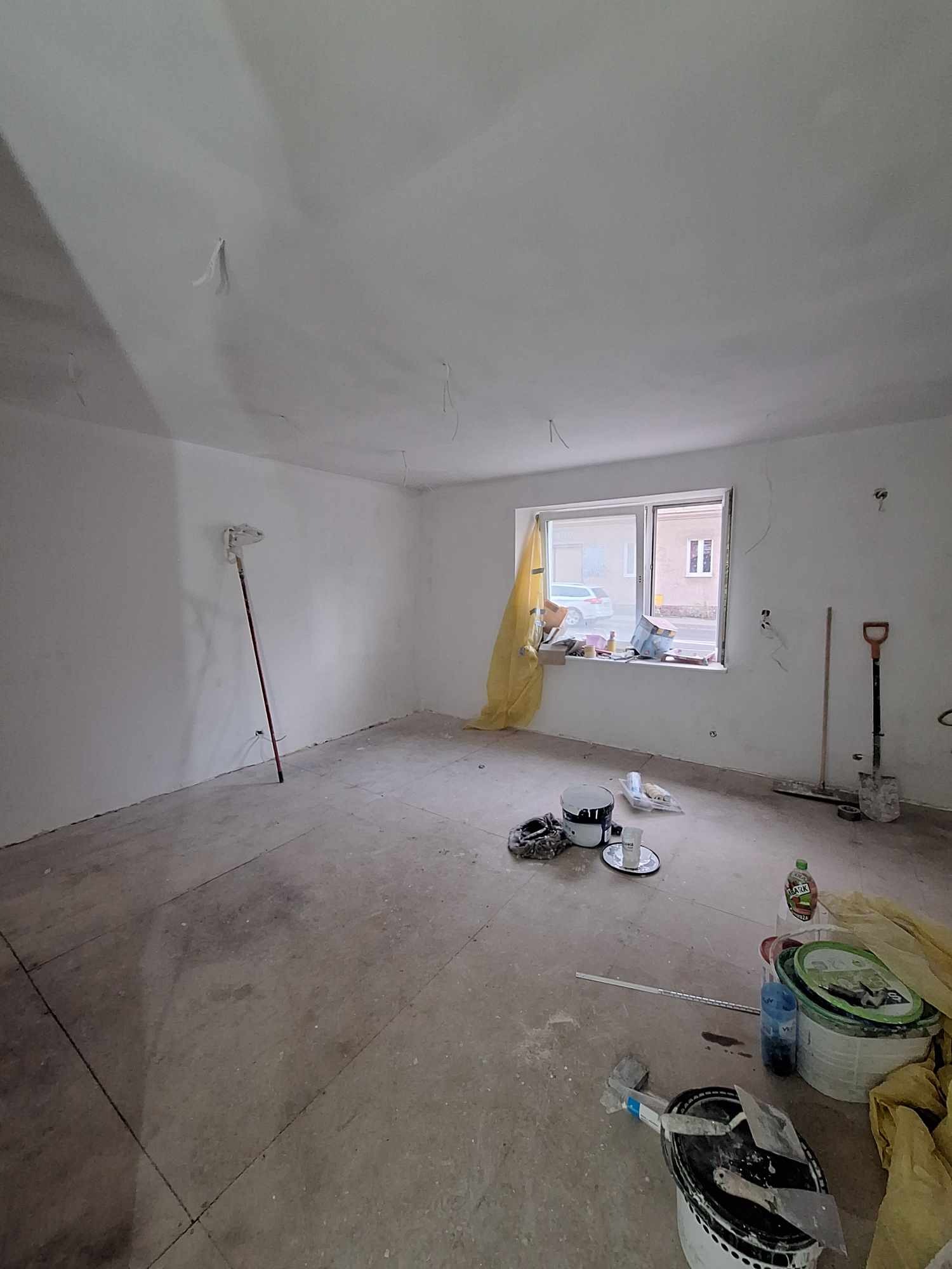 Pokój w trakcie remontu, widoczne ślady szpachlowania na suficie, narzędzia malarskie, wiaderka z farbą, zabezpieczone okno folią malarską.