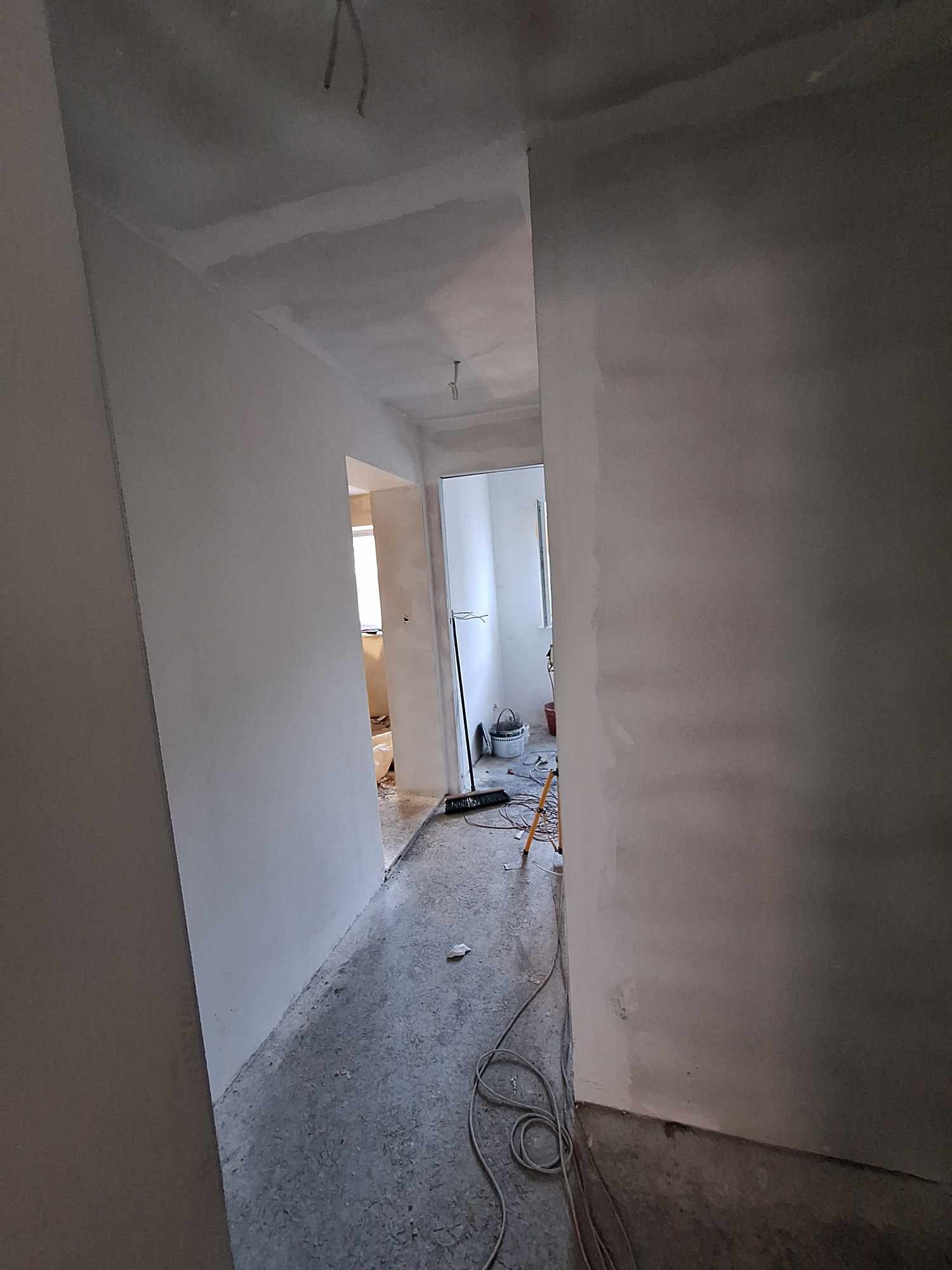 Wnętrze w trakcie prac wykończeniowych: świeżo wyszpachlowane ściany i sufit, widoczne narzędzia i wiadro w tle, przewody zwisające z sufitu.