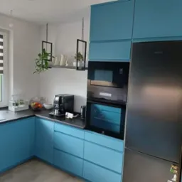 Turkusowa zabudowa kuchenna z czarnym blatem, piekarnikiem, mikrofalówką i lodówką, uzupełniona wiszącymi półkami z roślinami i oknem z żaluzjami.