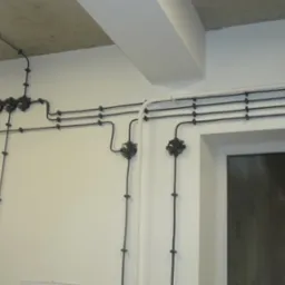 Czarna instalacja elektryczna natynkowa z puszkami rozgałęźnymi w stylu retro, zamontowana na białej ścianie w budynku w Łodzi, widoczna obok okna.