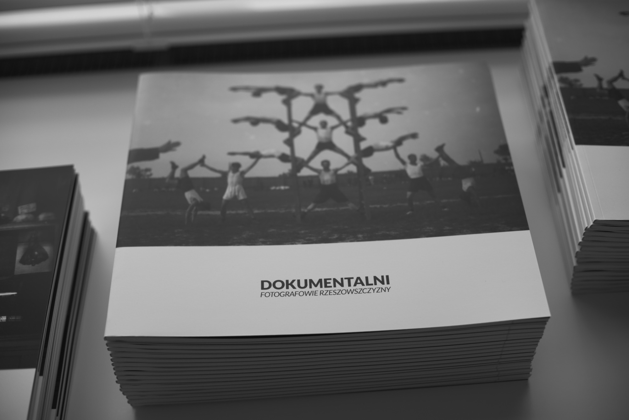 Stos czarno-białych albumów fotograficznych z napisem 'DOKUMENTALNI FOTOGRAFOWIE RZESZOWSZCZYZNY' i rozmytym zdjęciem akrobatów w tle.