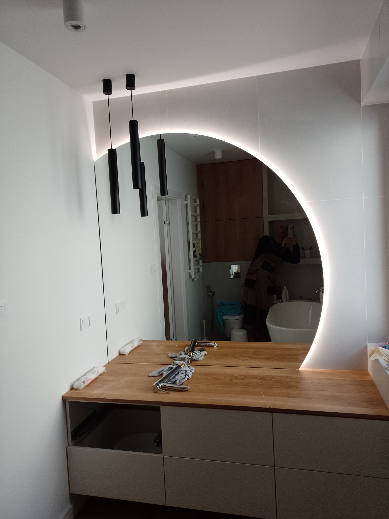 Łazienka w trakcie montażu: podświetlane lustro w kształcie półksiężyca nad drewnianym blatem z szufladami, widoczne narzędzia i armatura, odbicie osoby wykonującej zdjęcie.