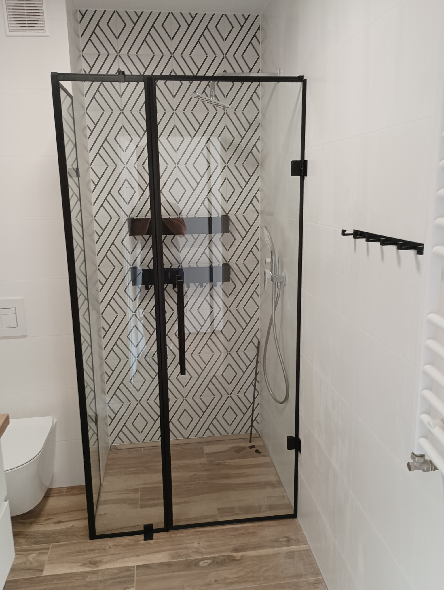 Nowoczesna kabina prysznicowa z czarnymi ramami i drzwiami składanymi, na tle białych płytek z geometrycznym wzorem, widoczny fragment toalety i grzejnika.