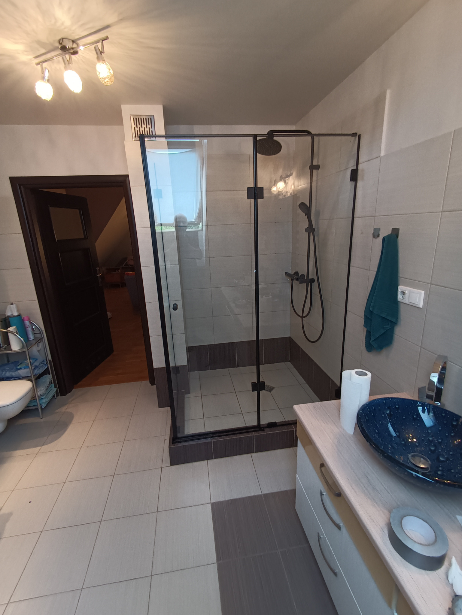 Nowoczesna kabina prysznicowa z czarnymi profilami i deszczownicą w łazience z beżowymi płytkami, widok na otwarte drzwi do innego pomieszczenia.