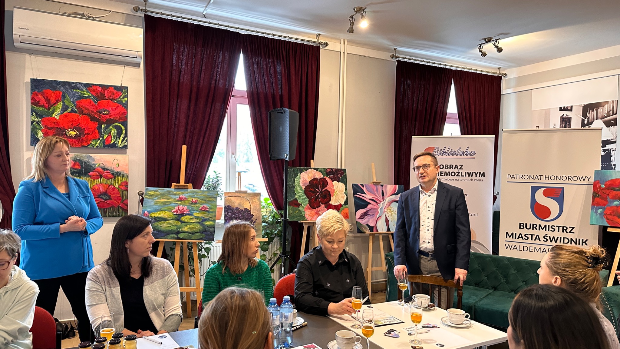 Prezentacja w pomieszczeniu z obrazami na sztalugach, uczestnicy siedzą przy stole z napojami, w tle banery 'Biblioteka' i 'Burmistrz Miasta Świdnik'.