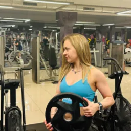 Kobieta w sportowym stroju trzyma czarny obciążnik 10 kg na tle siłowni z maszynami do ćwiczeń i innymi osobami trenującymi.