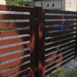 Nowoczesna brama wjazdowa z poziomych profili w kolorze brązowym, z czarną klamką, zamontowana przy ogrodzeniu z tego samego materiału, na tle trawnika i domu.