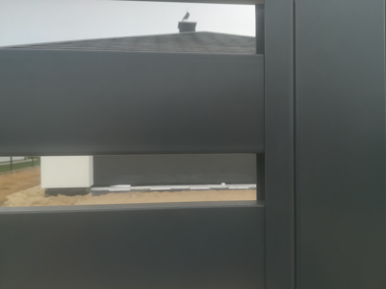 Fragment nowoczesnego, poziomego ogrodzenia w kolorze antracytowym, z widocznym w tle budynkiem i fragmentem trawnika.