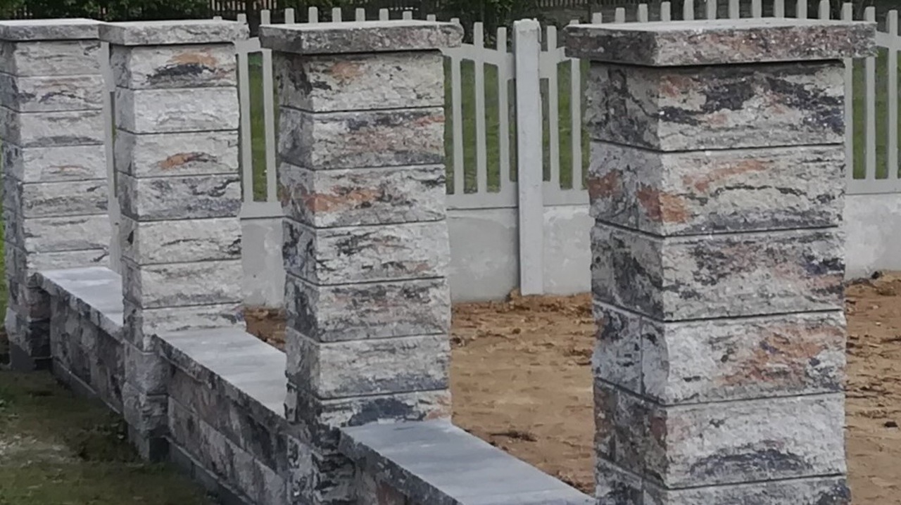 Fragment ogrodzenia z betonowych bloczków imitujących kamień, z widoczną szarą, betonową podmurówką i przęsłami ogrodzeniowymi w tle.