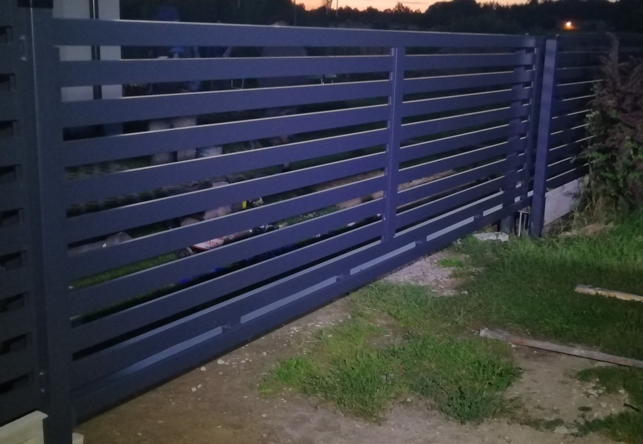 Nowoczesna, przesuwna brama w kolorze grafitowym z poziomymi panelami, widoczna z niskiej perspektywy na tle trawy i zarysów drzew o zmierzchu.