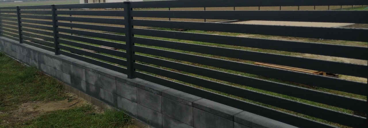 Nowoczesne, poziome ogrodzenie z szarych, metalowych paneli, osadzone na niskim murku z betonowych bloczków, z widoczną paletą w tle.