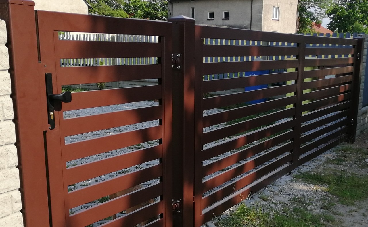 Nowoczesna brama wjazdowa z poziomych profili w kolorze brązowym, z czarną klamką, zamontowana przy ogrodzeniu z tego samego materiału, na tle trawnika i domu.