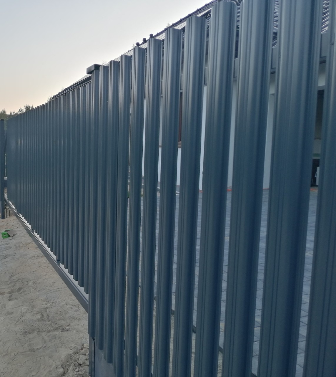 Nowoczesne, grafitowe ogrodzenie panelowe z pionowych, regularnych profili, widoczne z bliska pod kątem, z fragmentem zieleni w tle.