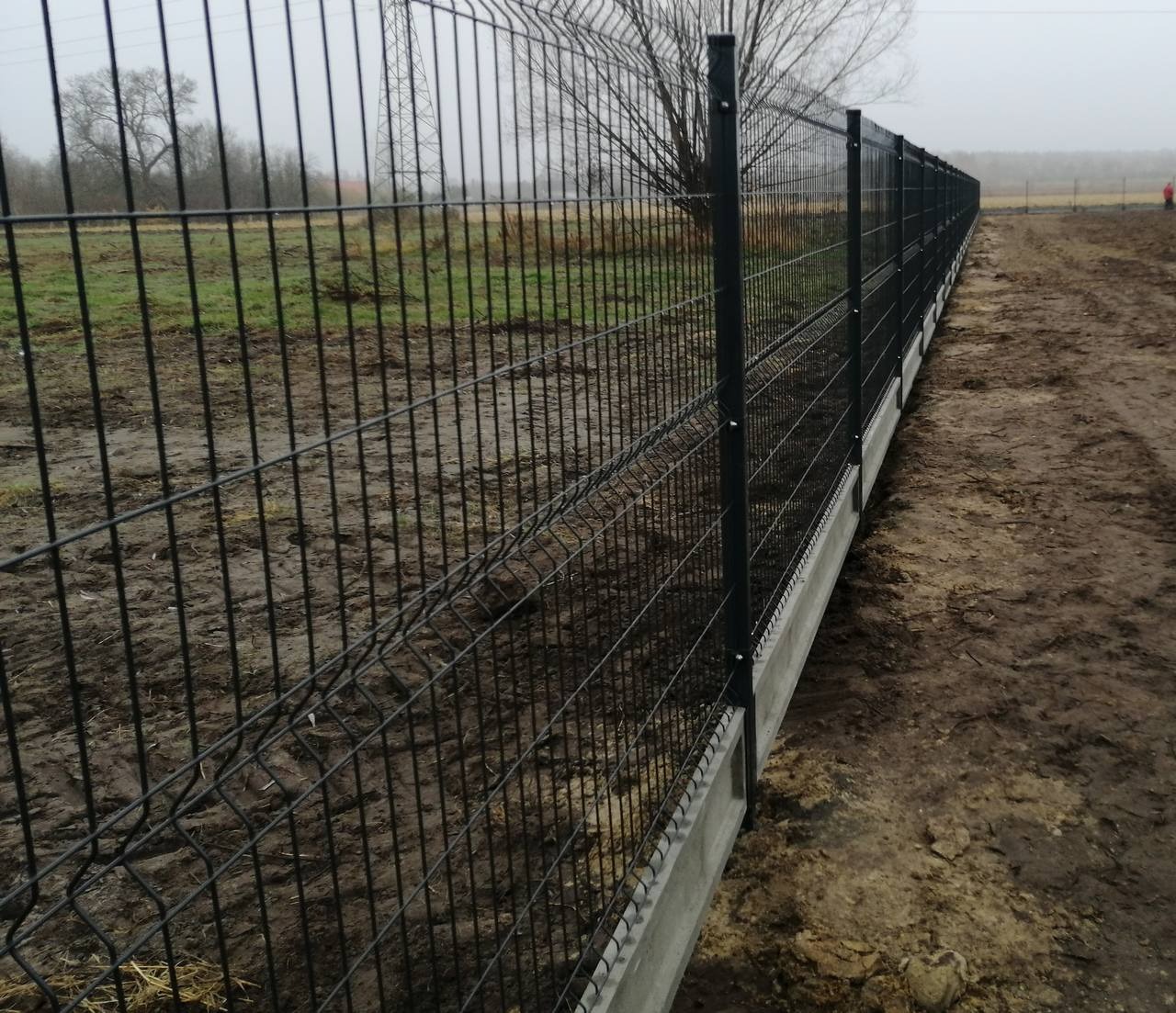 Długi, czarny panelowy płot z podmurówką betonową, rozciągający się wzdłuż błotnistego terenu, z widoczną postacią w oddali i słupami wysokiego napięcia w tle.