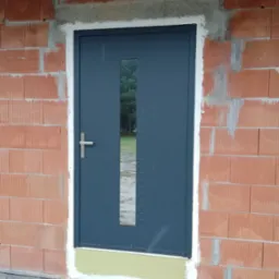 Szara, stalowa drzwi zewnętrzne z wąskim przeszkleniem pionowym, zamontowane w surowej ścianie z czerwonej cegły, widoczne ślady zaprawy i pianki montażowej.