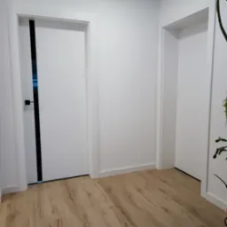 Nowoczesne białe drzwi wewnętrzne z czarnym akcentem, widoczne dwie pary drzwi w jasnym wnętrzu z drewnianą podłogą.