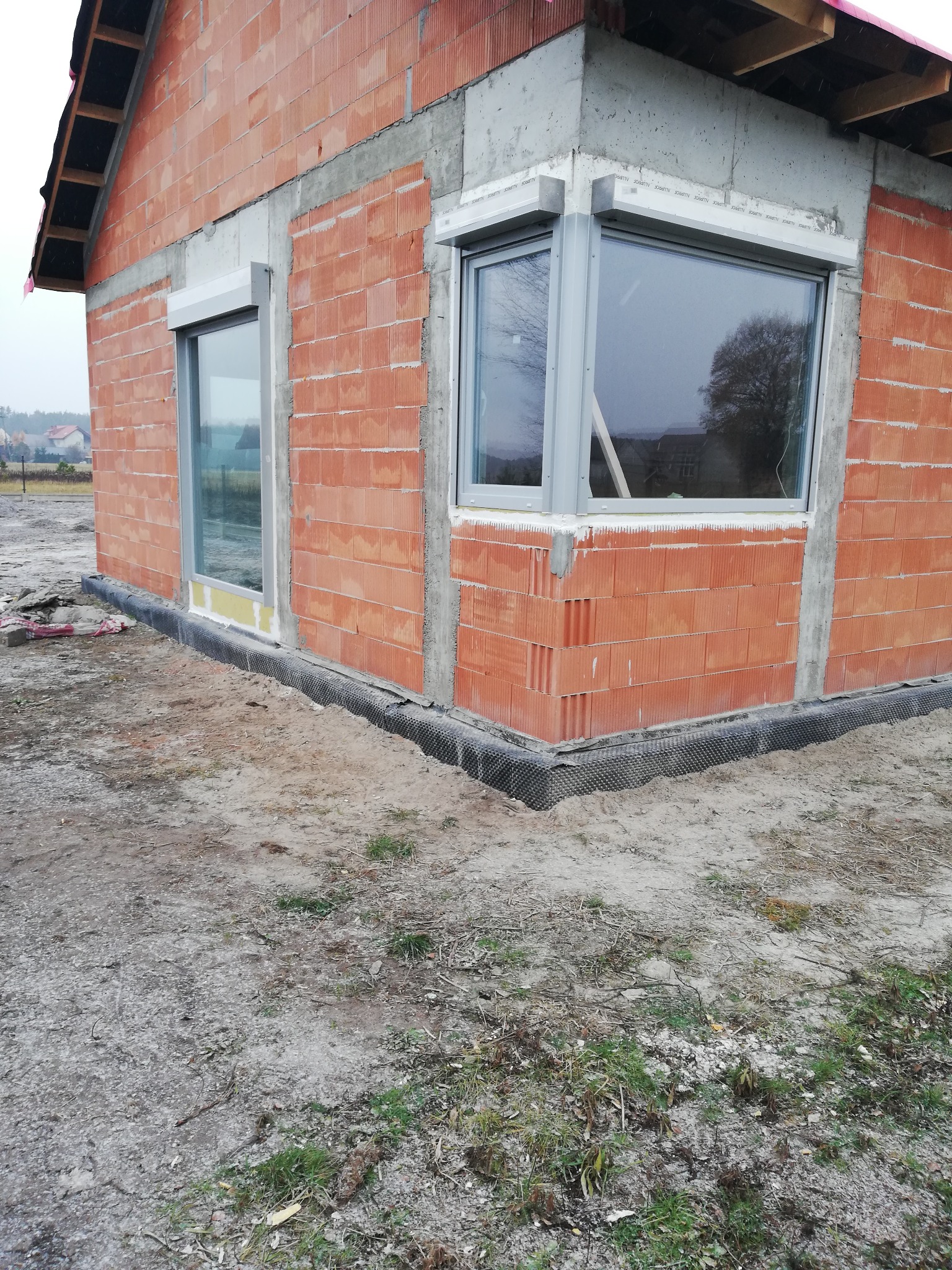 Surowy stan budynku z czerwonej cegły z zamontowanymi oknami PCV i roletami zewnętrznymi, widoczne szare ramy okienne i zwinięte rolety nad oknami, na dole budynku czarna folia izolacyjna.