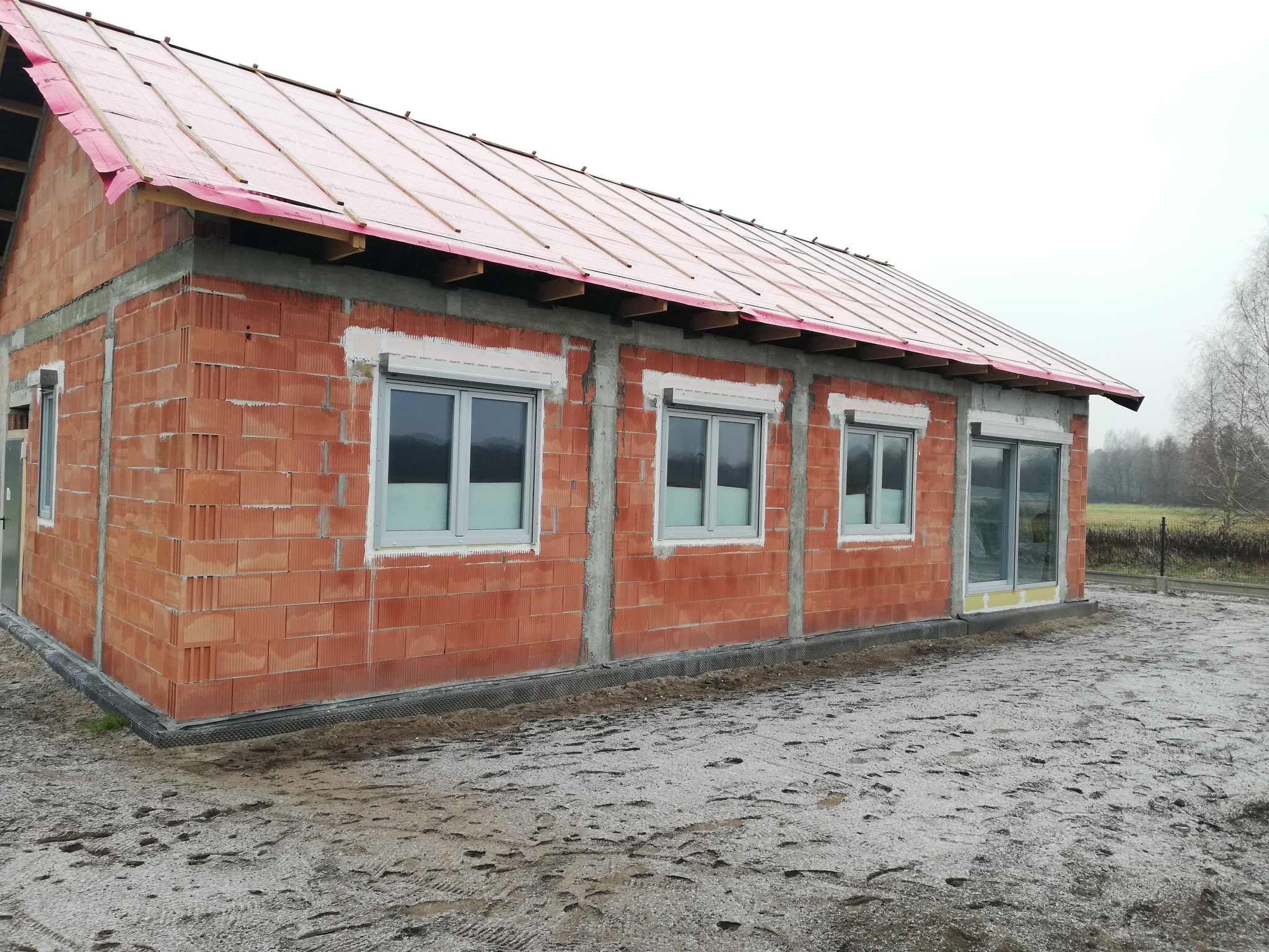 Surowy budynek z czerwonej cegły z zamontowanymi oknami PCV, w trakcie budowy dachu, widoczne elementy izolacji i konstrukcji, szare niebo w tle.