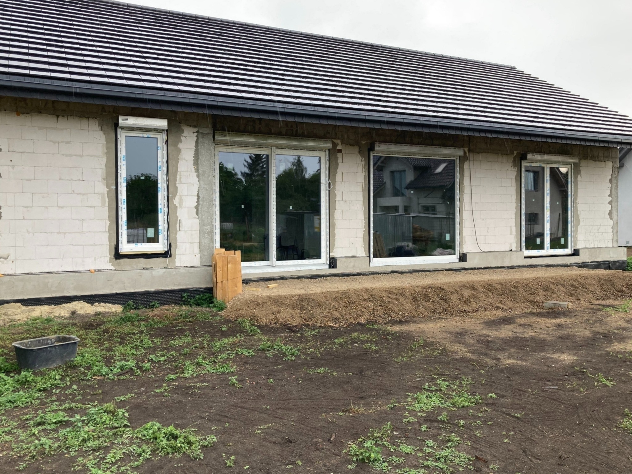 Dom w trakcie budowy z zamontowanymi oknami PCV z roletami zewnętrznymi, widoczne niedokończone elewacje i przygotowany teren wokół budynku.