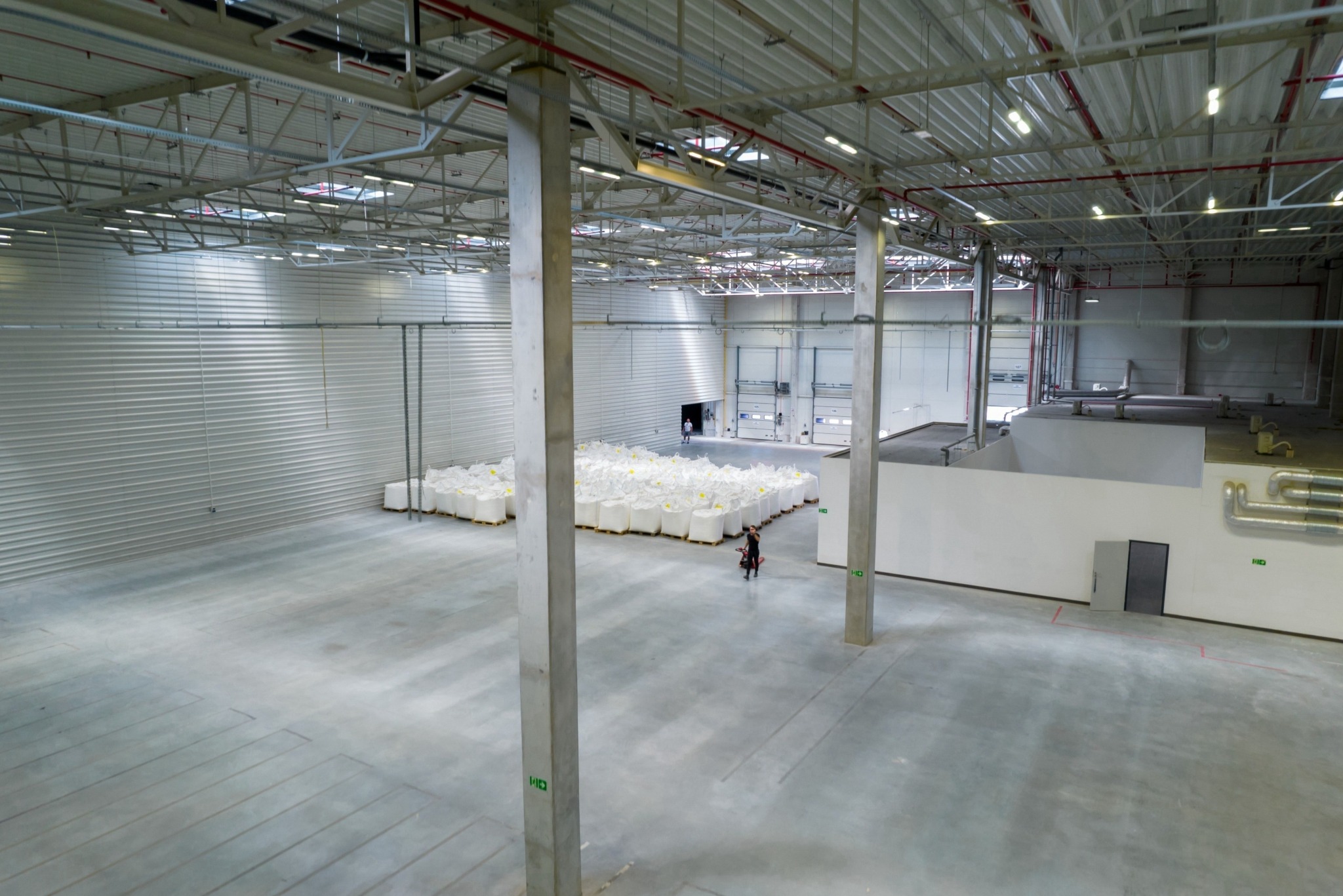Przestronny magazyn z rzędem białych worków ułożonych na paletach, widoczne betonowe filary, stalowa konstrukcja dachu i pracownik na wózku widłowym w oddali.