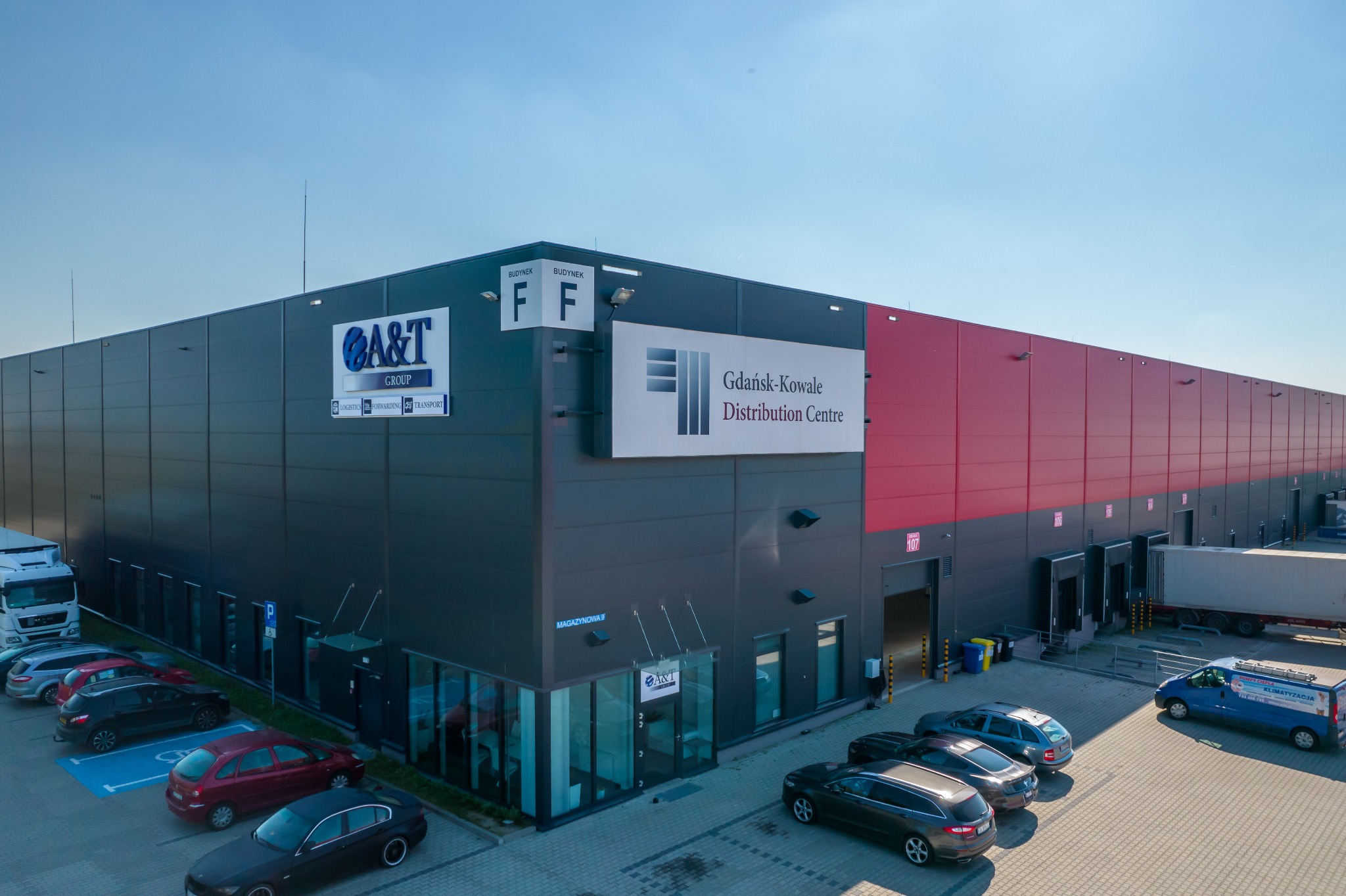 Widok z góry na nowoczesny budynek magazynowy Gdańsk-Kowale Distribution Centre z rampami załadunkowymi, parkingiem i oznakowaniem A&T Group w słoneczny dzień.