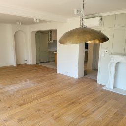 Usługi ogólno budowlane selen beata okrasa - Jasny parkiet w przestronnym wnętrzu po remoncie, widoczny fragment kuchni w odcieniach zieleni, biały kominek i złota lampa wisząca z folią ochronną na łańcuchu, świeżo pomalowane ściany.