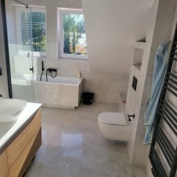 Usługi ogólno budowlane selen beata okrasa - Jasna łazienka na poddaszu z oknami, wanną, umywalką nablatową na drewnianej szafce, toaletą i grzejnikiem drabinkowym z powieszonym jasnoniebieskim ręcznikiem.