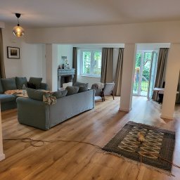 Usługi ogólno budowlane selen beata okrasa - Jasne, przestronne wnętrze salonu z drewnianą podłogą, szarymi sofami i dywanem w etniczne wzory, widok na ogród przez przeszklone drzwi balkonowe.