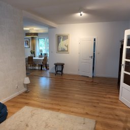 Usługi ogólno budowlane selen beata okrasa - Jasne panele podłogowe w salonie z widokiem na jadalnię, wzorzysta tapeta na ścianie, dekoracyjny obraz na ścianie, białe drzwi, mały stolik i lampa podłogowa.