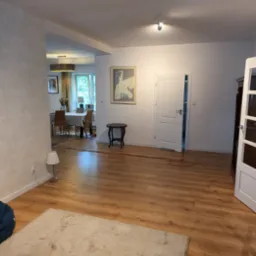 Jasne panele podłogowe w salonie z widokiem na jadalnię, wzorzysta tapeta na ścianie, dekoracyjny obraz na ścianie, białe drzwi, mały stolik i lampa podłogowa.