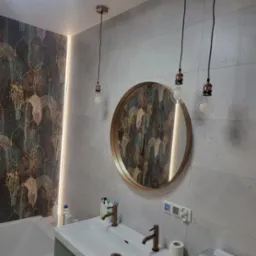 Nowoczesna łazienka z umywalką z dwoma kranami w kolorze miedzi, okrągłym lustrem w złotej ramie, trzema wiszącymi lampami z żarówkami Edisona i płytkami z motywem liści.