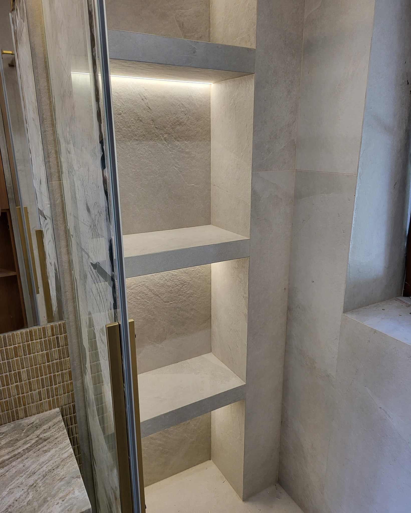 Wykończona wnęka z półkami podświetlonymi taśmą LED, obok kabina prysznicowa z złotymi uchwytami, całość wyłożona płytkami imitującymi kamień.
