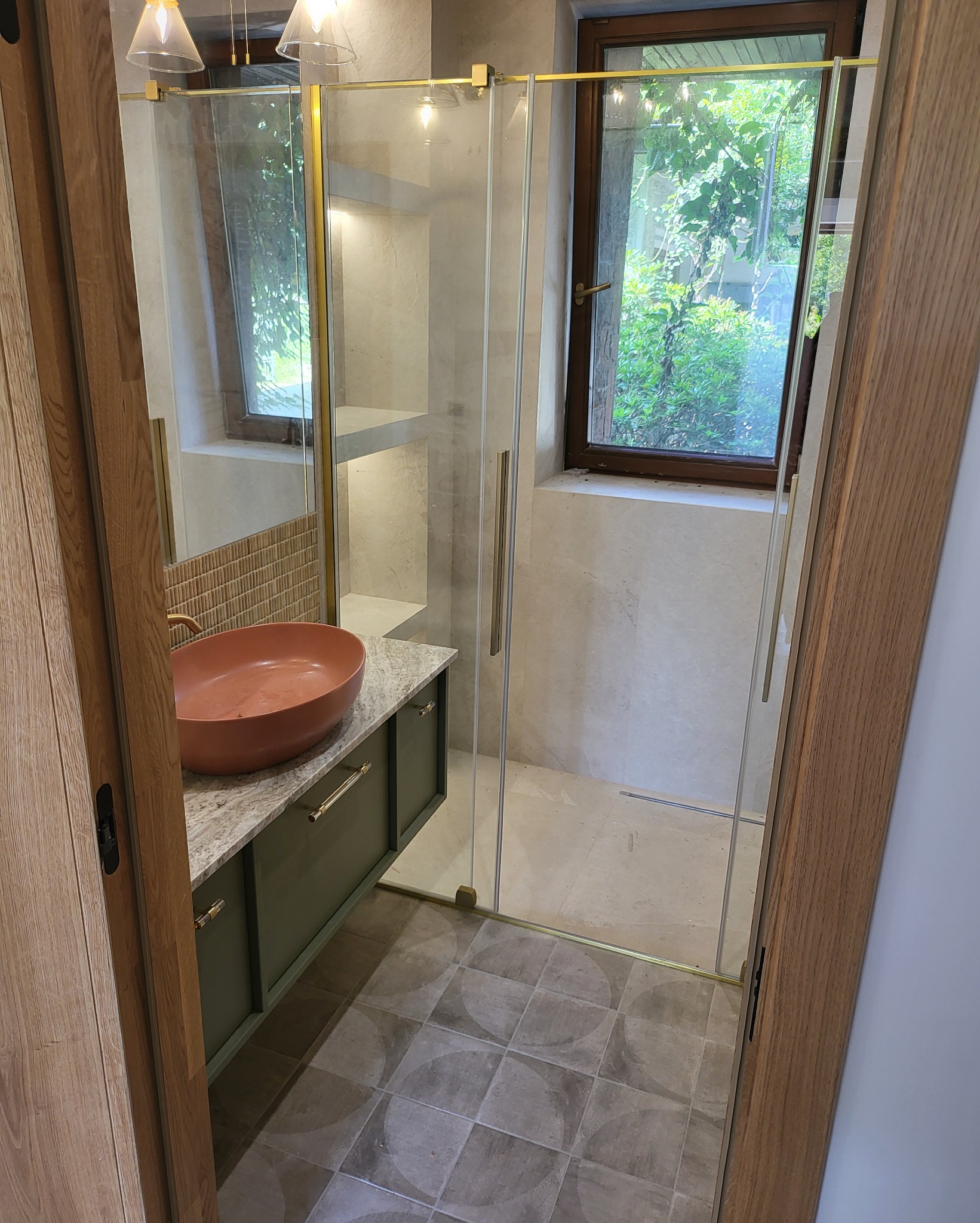 Nowoczesna łazienka z kabiną prysznicową ze złotymi elementami, umywalką w kolorze terakoty na szafce z blatem imitującym marmur, widok przez drzwi z drewnianą ramą