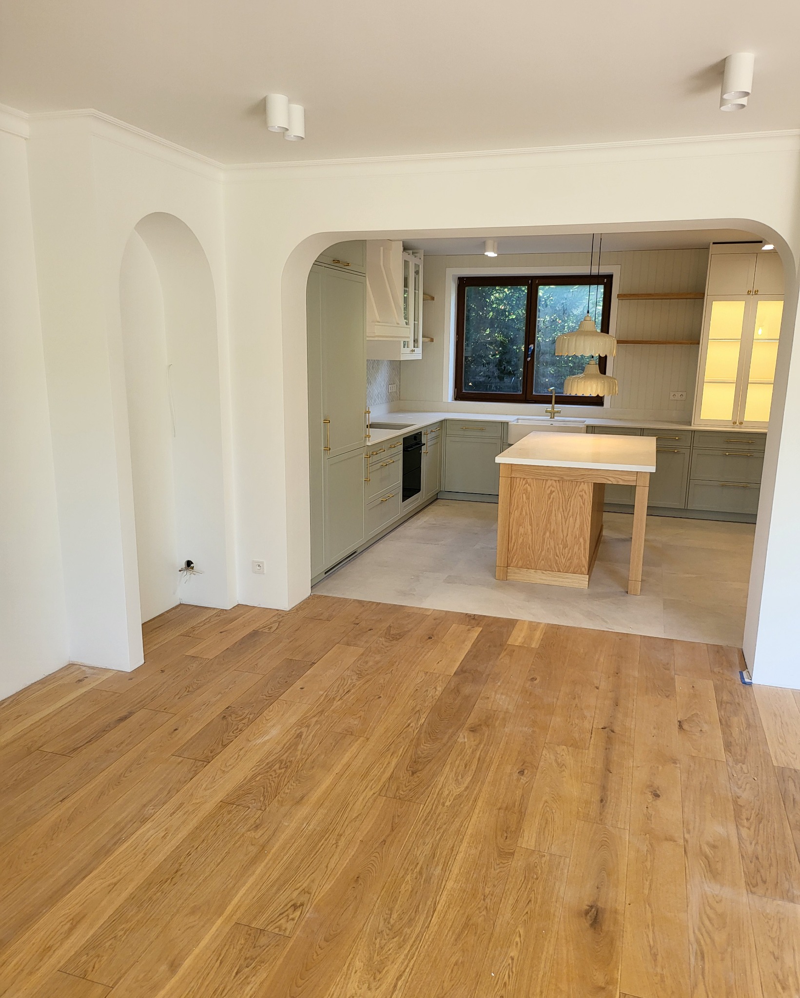 Jasna, przestronna kuchnia z drewnianą wyspą i pastelowymi szafkami, widok z salonu z drewnianą podłogą i łukowym przejściem.