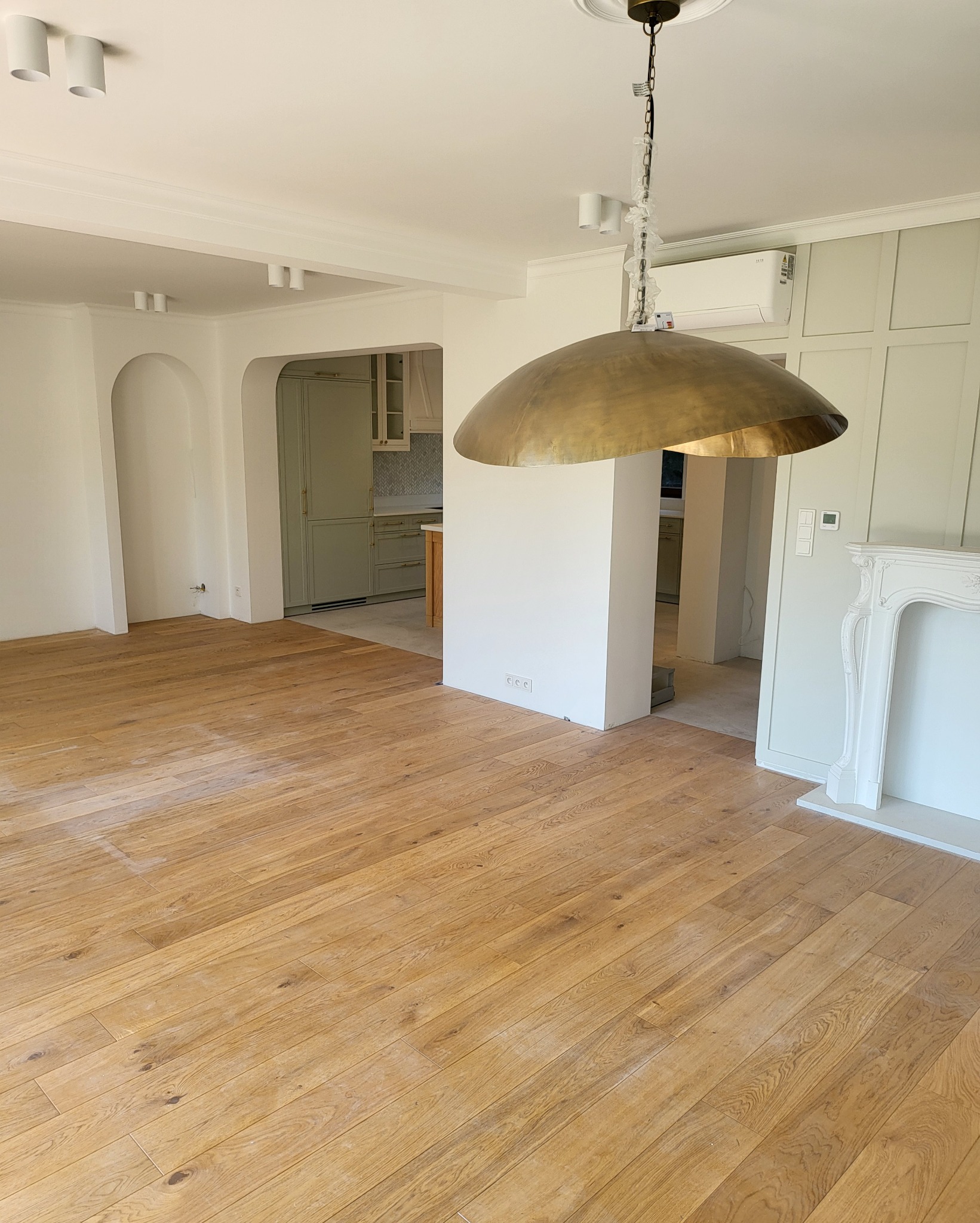 Jasny parkiet w przestronnym wnętrzu po remoncie, widoczny fragment kuchni w odcieniach zieleni, biały kominek i złota lampa wisząca z folią ochronną na łańcuchu, świeżo pomalowane ściany.