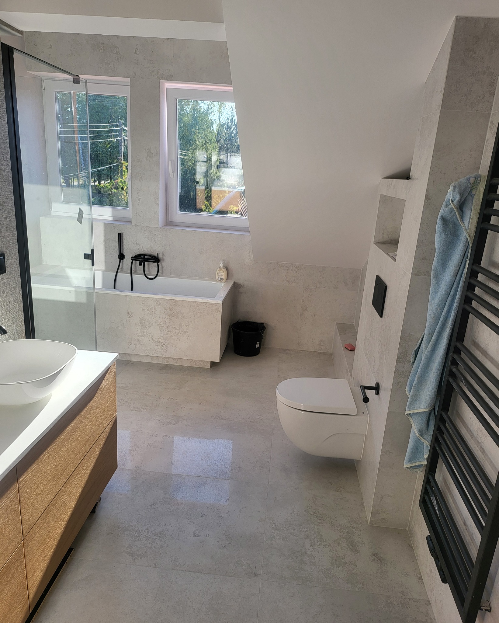 Jasna łazienka na poddaszu z oknami, wanną, umywalką nablatową na drewnianej szafce, toaletą i grzejnikiem drabinkowym z powieszonym jasnoniebieskim ręcznikiem.