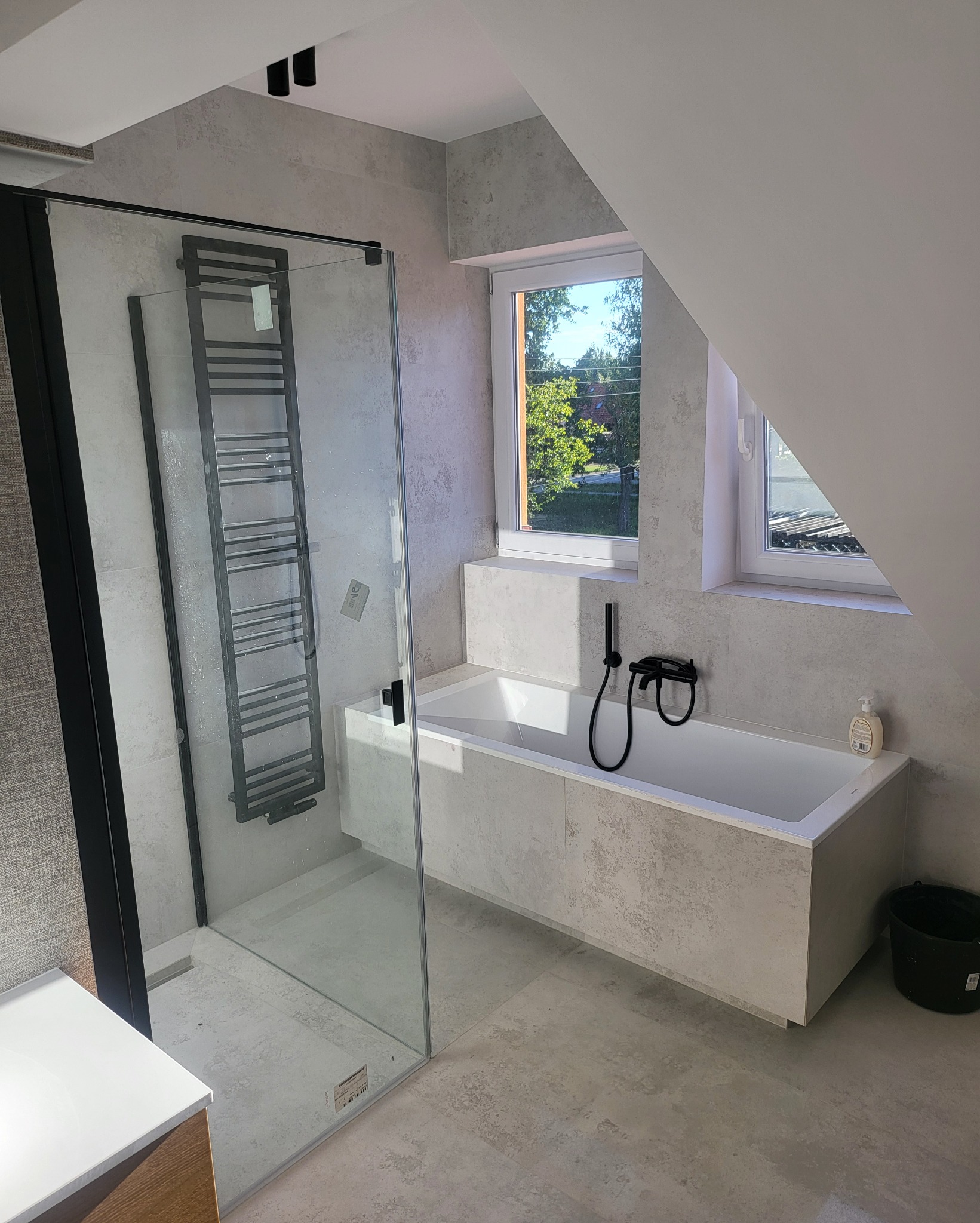 Nowoczesna łazienka na poddaszu z kabiną prysznicową typu walk-in, grzejnikiem drabinkowym, wanną obudowaną płytkami imitującymi beton, czarną armaturą i oknami dachowymi.