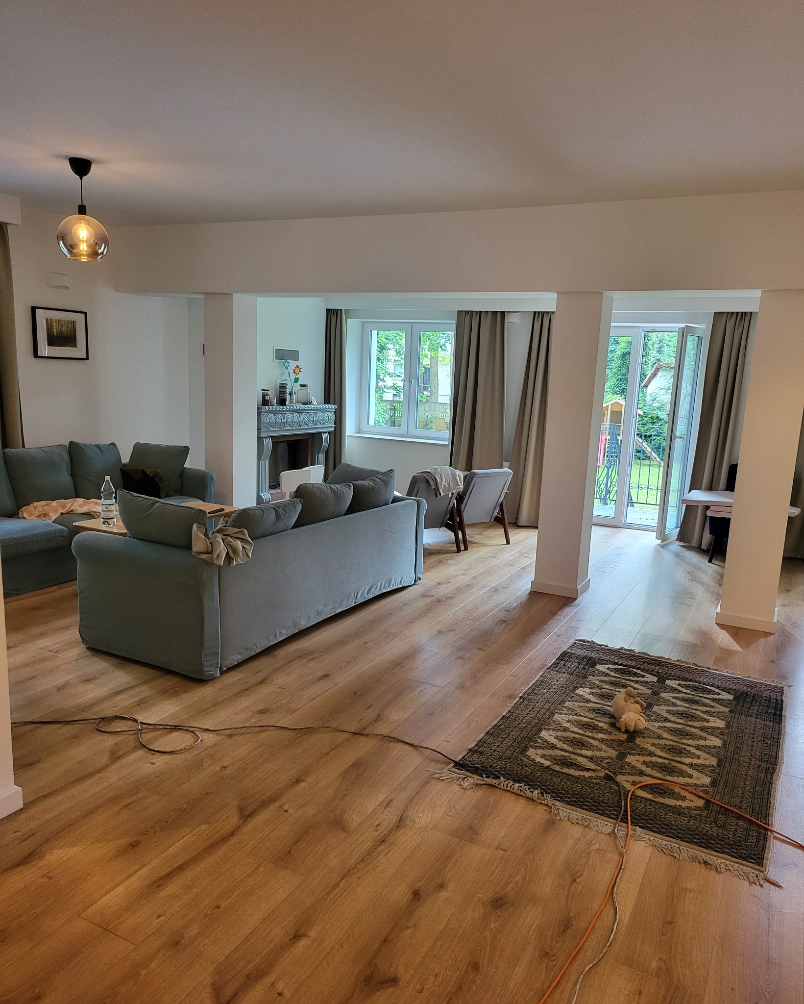 Jasne, przestronne wnętrze salonu z drewnianą podłogą, szarymi sofami i dywanem w etniczne wzory, widok na ogród przez przeszklone drzwi balkonowe.