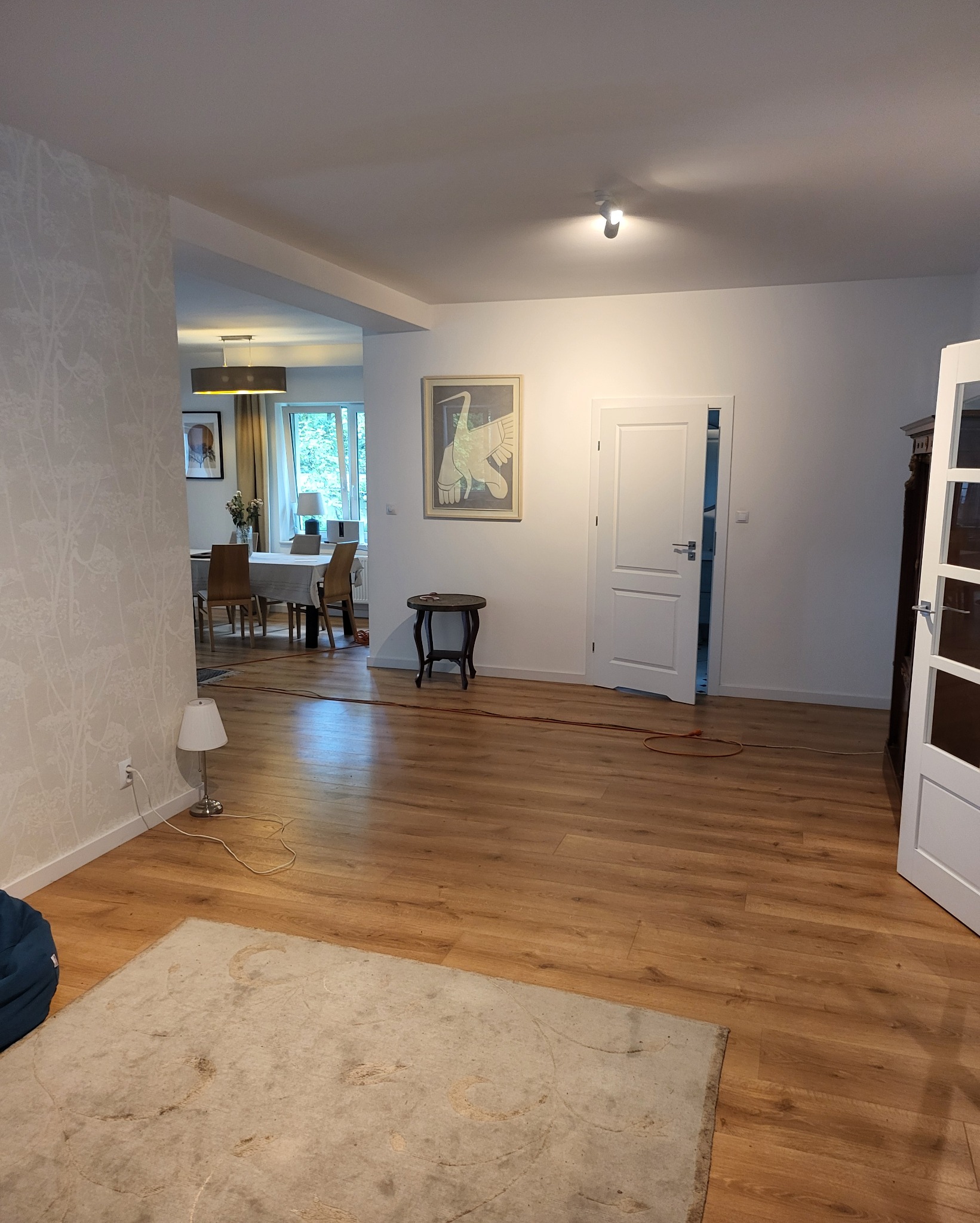 Jasne panele podłogowe w salonie z widokiem na jadalnię, wzorzysta tapeta na ścianie, dekoracyjny obraz na ścianie, białe drzwi, mały stolik i lampa podłogowa.