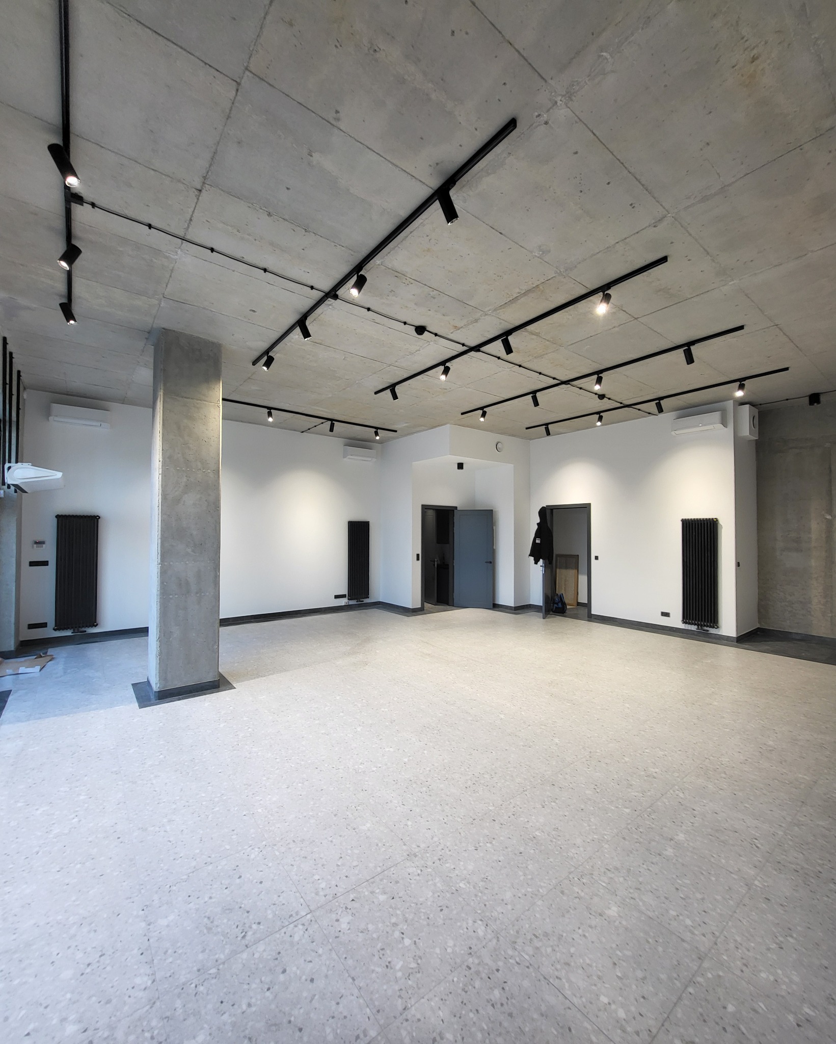 Przestronne, jasne wnętrze z betonowym sufitem, czarnymi reflektorami szynowymi, białymi ścianami, szarą posadzką lastryko, czarnymi grzejnikami i betonowym słupem.